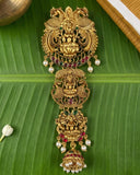 An ornate gold-toned traditional Indian jewelry piece with intricate filigree work, deity motifs, red and green stone accents, and pearl-like white beads, displayed on a large green banana leaf.