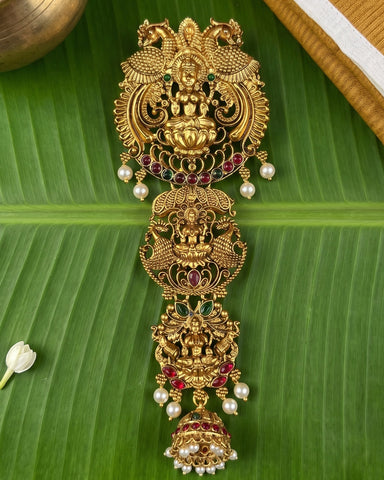 An ornate gold-toned traditional Indian jewelry piece with intricate filigree work, deity motifs, red and green stone accents, and pearl-like white beads, displayed on a large green banana leaf.