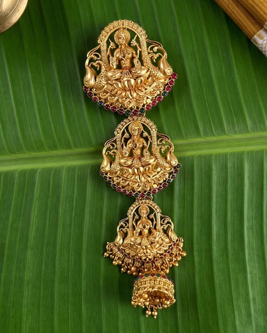 An ornate gold-toned traditional Indian jewelry piece featuring three stacked deity motifs with red gemstone accents, displayed on a green banana leaf.