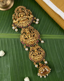 An ornate gold-toned traditional Indian jewelry piece with intricate filigree, deity carvings, and dangling pearl-like beads, arranged on a green banana leaf with small white flower petals and a brass object at the corner.