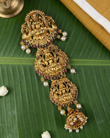 An ornate gold-toned traditional Indian jewelry piece with intricate filigree, deity carvings, and dangling pearl-like beads, arranged on a green banana leaf with small white flower petals and a brass object at the corner.
