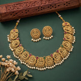 Gold-plated Diwali necklace and matching earrings with intricate filigree, decorated with white pearls, red and green stone accents, and displayed on a dark green background with a carved wooden prop.