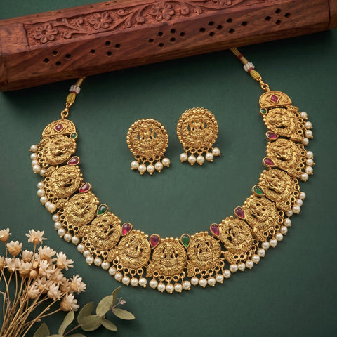 Gold-plated Diwali necklace and matching earrings with intricate filigree, decorated with white pearls, red and green stone accents, and displayed on a dark green background with a carved wooden prop.