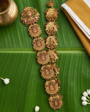 An ornate gold-toned traditional necklace with multiple circular and floral motifs, decorated with red and green stones, displayed on a large green banana leaf with small white flowers and a brass pot in the background.