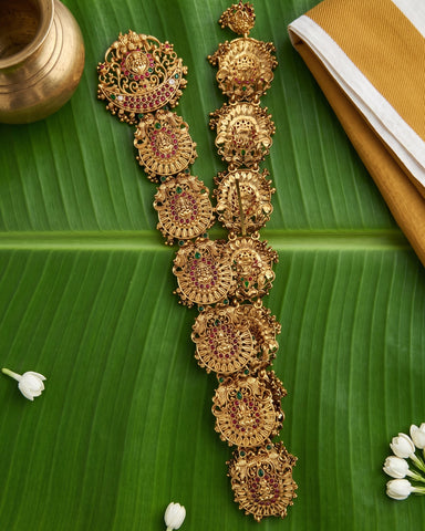 An ornate gold-toned traditional necklace with multiple circular and floral motifs, decorated with red and green stones, displayed on a large green banana leaf with small white flowers and a brass pot in the background.