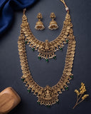An ornate gold-toned traditional necklace set with matching earrings, decorated with coin-like motifs, small white pearl-like beads, and green jewel accents, arranged on a dark navy background with decorative fabric and dried flowers.