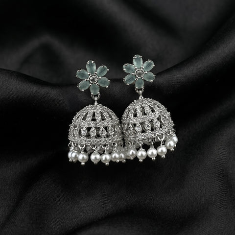 A pair of silver-toned jhumka earrings with light blue-green flower-shaped studs and dangling pearl-like beads, displayed on a draped black satin fabric background.