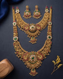 An ornate gold-toned jewelry set laid flat on a dark navy background, including a large necklace, matching earrings, and smaller matching pieces. The jewelry is decorated with white pearl-like stones, green emerald-cut stones, and red stone accents.