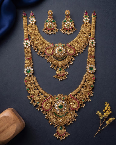 An ornate gold-toned jewelry set laid flat on a dark navy background, including a large necklace, matching earrings, and smaller matching pieces. The jewelry is decorated with white pearl-like stones, green emerald-cut stones, and red stone accents.