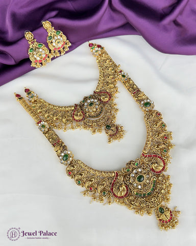 Ornate gold necklace with green and red stones on a white surface with purple fabric in the background, featuring 'Jewel Palace' branding.