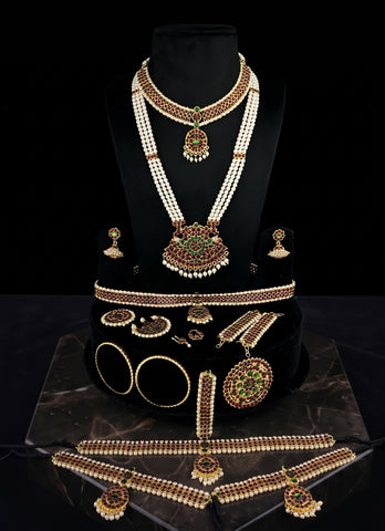 A traditional Indian gold and white pearl jewelry set displayed on a black mannequin bust and stand. The set includes a multi-strand pearl necklace with a large green and gold pendant, matching earrings, a bracelet, a waist belt, and other coordinating jewelry pieces.