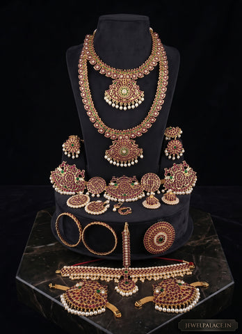 An ornate Indian bridal jewelry set displayed on a black mannequin bust and marble pedestal against a black background. The set includes a gold-toned necklace with red and green stone embellishments and white pearl bead borders, matching earrings, a bracelet, a necklace holder, and a long pendant.