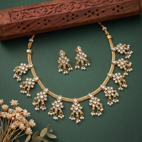 Gold-plated necklace and matching earrings with white pearl-like accents, arranged on a dark green background with decorative dried flowers and a carved wooden prop.
