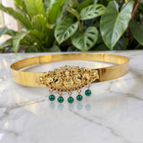A gold-toned traditional Indian choker necklace with an ornate central filigree piece featuring Ganesha and peacock motifs, accented with small red stones and three dangling green pearl beads, displayed on a white marble surface with green plants in the background.