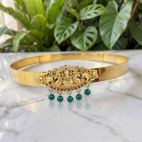 A gold-toned traditional Indian choker necklace with an ornate central filigree piece featuring Ganesha and peacock motifs, accented with small red stones and three dangling green pearl beads, displayed on a white marble surface with green plants in the background.