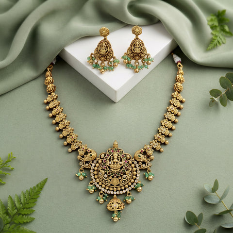 An ornate gold-toned traditional necklace and matching dangle earrings displayed on a white rectangular block, with draped sage green fabric and scattered green leaves around them.