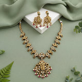 Gold-toned traditional necklace and matching earrings with green and pink stone accents, arranged on a sage green fabric background with a white display block and small green leaves.