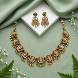 Gold-toned traditional jewelry set with a necklace and matching earrings, decorated with gold-toned motifs, white stones, and red and green gem accents, arranged on a sage green draped background with a white display block and small white flowers.