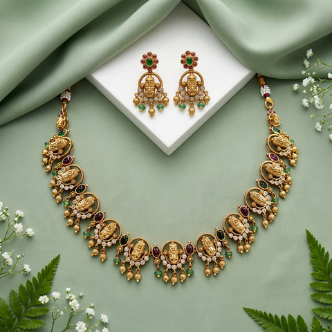 Gold-toned traditional jewelry set with a necklace and matching earrings, decorated with gold-toned motifs, white stones, and red and green gem accents, arranged on a sage green draped background with a white display block and small white flowers.
