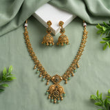Gold-toned traditional Indian jewelry set featuring a necklace and matching earrings with intricate filigree work, small green bead accents, and a central paisley pendant, arranged on a sage green fabric backdrop with a white block and decorative leaves.