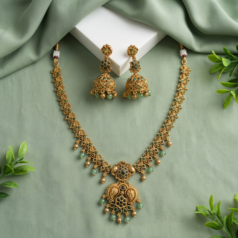 Gold-toned traditional Indian jewelry set featuring a necklace and matching earrings with intricate filigree work, small green bead accents, and a central paisley pendant, arranged on a sage green fabric backdrop with a white block and decorative leaves.