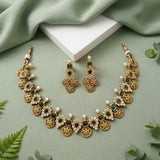 Gold-toned traditional jewelry set with a necklace and matching earrings, decorated with green rectangular stones, white faux pearls, and floral motifs, arranged on a sage green fabric background with a white block and small green leaves.