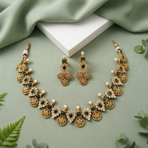 Gold-toned traditional jewelry set with a necklace and matching earrings, decorated with green rectangular stones, white faux pearls, and floral motifs, arranged on a sage green fabric background with a white block and small green leaves.