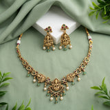 Gold-toned Indian jewelry set featuring a necklace and matching earrings with detailed deity motifs, pearl-like white beads, and small green stone accents, arranged on a sage green draped fabric background with a white display block and leaf props.