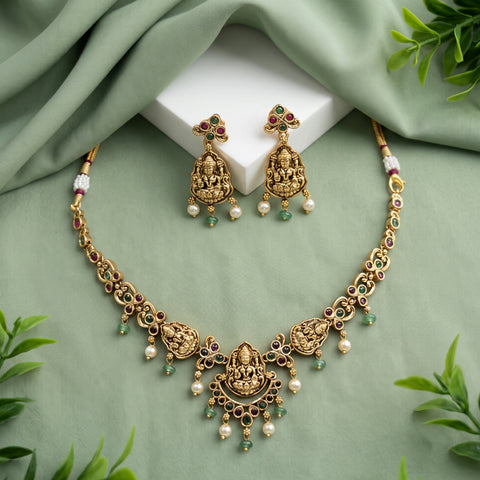 Gold-toned Indian jewelry set featuring a necklace and matching earrings with detailed deity motifs, pearl-like white beads, and small green stone accents, arranged on a sage green draped fabric background with a white display block and leaf props.