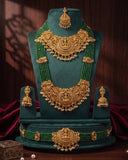 A traditional Indian gold jewelry set displayed on a dark green velvet stand, featuring a necklace, matching earrings, a waist chain, and a maang tikka. The jewelry is detailed with intricate gold filigree and adorned with green beads, set against a dark wood background with red drapery.