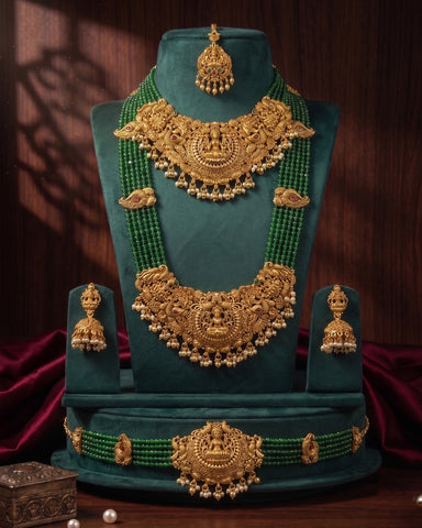 A traditional Indian gold jewelry set displayed on a dark green velvet stand, featuring a necklace, matching earrings, a waist chain, and a maang tikka. The jewelry is detailed with intricate gold filigree and adorned with green beads, set against a dark wood background with red drapery.