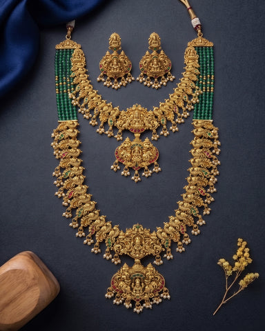 An ornate gold-toned traditional necklace set with matching earrings, decorated with green bead strands and small red and green stone accents, arranged on a dark navy background with decorative fabric and a wooden block.