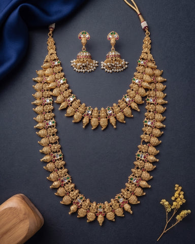 An ornate gold-toned traditional jewelry set featuring a double-strand choker necklace and matching jhumka earrings, decorated with intricate filigree, beaded details, and small red and green stone accents, arranged on a dark textured background with decorative fabric and a wooden block.
