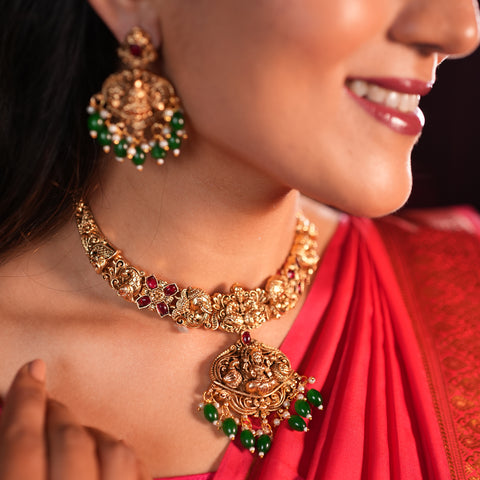 Close-up of a woman wearing gold jewelry with red and green stones, including a necklace and earrings, against a dark background.