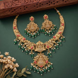 An ornate gold-toned traditional necklace and matching earrings with green bead drops, coral-red stone accents, and small white pearl-like beads, arranged on a dark green background with decorative dried flowers and a carved wooden prop.