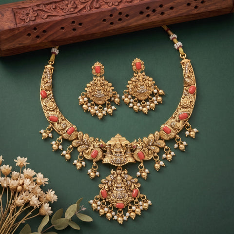 Gold-toned traditional Indian jewelry set featuring a statement necklace and matching earrings with coral-red stone accents and white pearl bead drops, arranged on a dark green background with decorative dried flowers and a carved wooden prop.