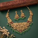 An ornate gold-toned traditional necklace with matching earrings, decorated with green bead drops, red floral accents, and small pearl-like beads, arranged on a dark green background with a carved wooden prop and dried flowers.