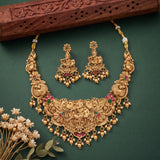 An ornate gold-toned traditional necklace set with matching earrings, decorated with floral and animal motifs and accented with red and green stones and white pearl-like beads, arranged on a dark green background with a carved wooden prop and small dried flowers.