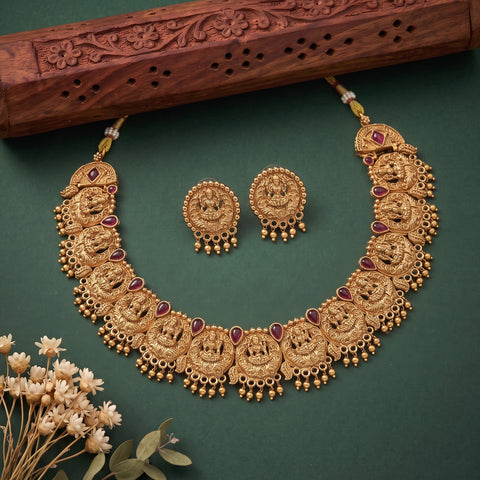 An ornate gold-toned traditional necklace set with matching earrings, decorated with red gemstone accents and dangling bead chains, arranged on a dark green background with a carved wooden prop and small dried flowers.