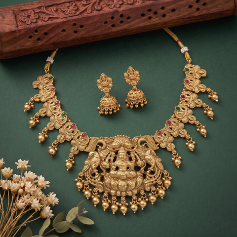 An ornate gold-toned traditional necklace set with matching earrings, displayed on a dark green background with dried flowers and a carved wooden prop. The necklace features detailed filigree, hanging gold beads, and small red and green stone accents.