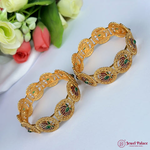 Gold bangles with intricate designs on a white surface with flowers in the background, brand 'Jewel Palace' visible.