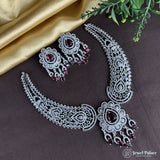 Silver necklace and earrings with red gemstones on a dark surface with green leaves.