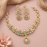Gold-toned traditional jewelry set with a necklace and matching earrings, decorated with clear stones and mint green stones, arranged on a dusty pink background with burlap and a pink flower prop.