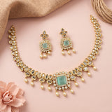 Gold-toned traditional jewelry set featuring a necklace and matching earrings with mint green rectangular stones, clear stones, and pearl-like drops, arranged on a soft pink background with decorative burlap and a pink flower.