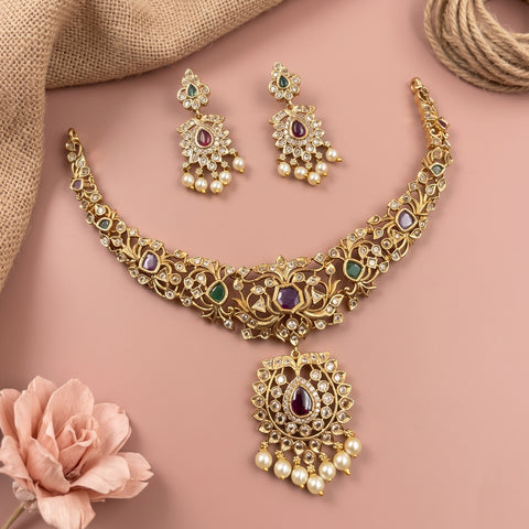Gold-toned Indian jewelry set featuring a necklace with a matching pendant and a pair of earrings, decorated with green and purple stones and white faux pearls, arranged on a pink background with decorative fabric and a flower.