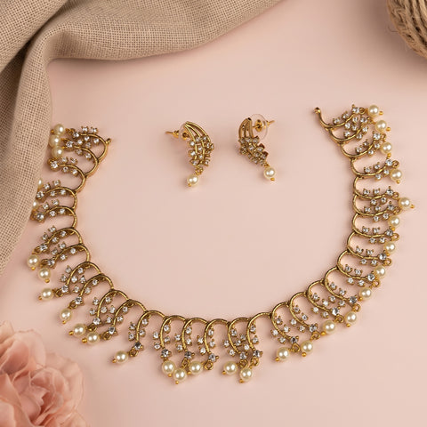 Gold-tone jewelry set featuring a statement collar necklace, matching drop earrings, and pearl accents, arranged on a soft pink background with a beige fabric and rose prop.