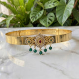 Gold-plated Indian choker necklace with a central ornate filigree piece featuring a red ruby and white stones, accented with green emeralds, and three dangling green bead drops, displayed on a white marble surface with green plants in the background.