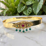Gold-plated choker necklace with an ornate central filigree design, a red teardrop gemstone, and five dangling green bead drops, displayed on a white marble surface with green plants in the background.