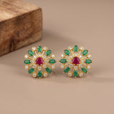 A pair of gold-toned floral stud earrings with green marquise-cut stones, clear round stones, and a central red oval stone, displayed on a beige surface with a wooden block in the background.