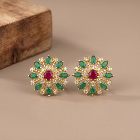 A pair of gold-toned floral stud earrings with green marquise-cut stones, clear round stones, and a central red oval stone, displayed on a beige surface with a wooden block in the background.